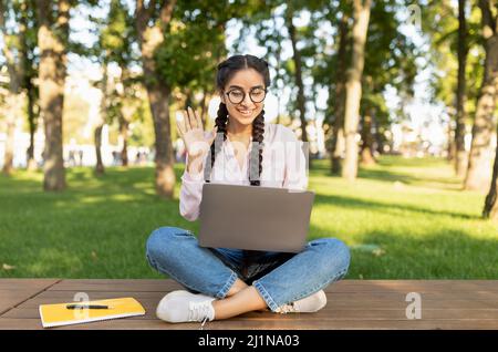 E-Learning. Glückliche Studentin mit Laptop, die an einem Online-Vortrag teilnimmt, die Hand an der Webcam schwenkt und draußen auf der Bank sitzt Stockfoto