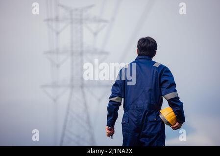 Ein männlicher Energietechniker mit gelbem Helm inspiziert die Stromleitung unter einer Hochspannungsleitung. Stockfoto