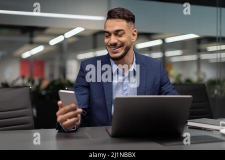 Lächelnder arabischer Geschäftsmann überprüft während der Arbeit die mobile App Stockfoto
