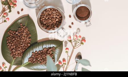 Geröstete Bio-Kaffeebohnen auf grünen Blättern und Kaffeepulver in einem Glasgefäß auf elfenbeinfarbenem Hintergrund mit Schatten und Kopierraum. Kaffeemaschinen konz Stockfoto