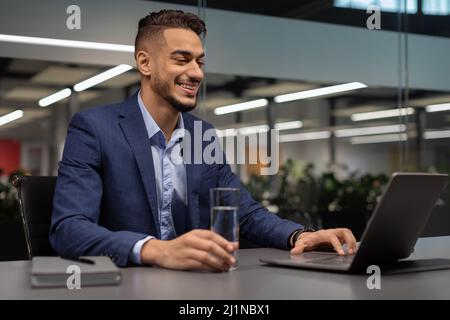 Glücklicher Manager, der Wasser trinkt, während er an einem Laptop arbeitet Stockfoto