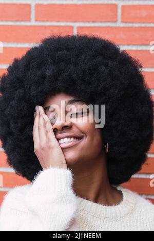 Porträt einer fröhlichen afroamerikanischen Frau mit weißem, perfektem Lächeln, langen Afro-Haaren und weißem Pullover, der das Auge an der Ziegelwand bedeckt. Zahnarztkonzept Stockfoto