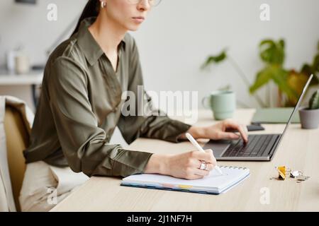 Seitenansicht Porträt einer Geschäftsbesitzerin, die im Notebook schreibt, während sie den Computer in minimalem Büroinnenraum benutzt Stockfoto