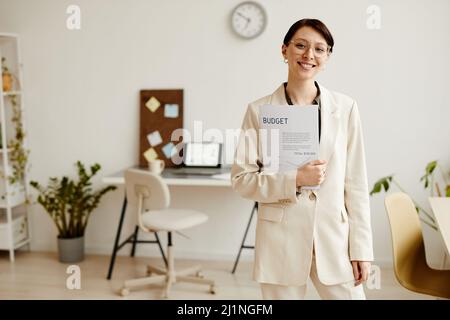 Porträt einer erfolgreichen jungen Geschäftsfrau, die mit einem Lächeln auf die Kamera lächelt, während sie in einem minimalen Büro steht und Platz für Kopien hat Stockfoto