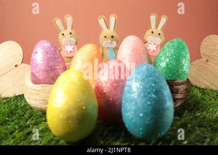 Drei osterhasen, die sich gleichmäßig hinter bunten ostereiern auf einem orange-rötlichen Hintergrund mit Holzherzen auf beiden Seiten verteilt befinden. Stockfoto