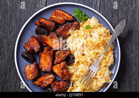 Geschmortes Sauerkraut mit Schweinebraten, kurze Rippen und Pflaumen auf Teller auf dunklem Holztisch, horizontale Ansicht von oben, flaches Lay, Makro Stockfoto