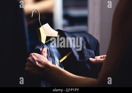 Mann, der sich fertig gemacht hat und sich in einem Anzug kleidet. Luxuriöse Jacke und Krawatte im Kleiderbügel. Versuchen Sie auf Outfit vor Datum oder Hochzeit. Umkleidezimmer im Einkaufszentrum. Stockfoto