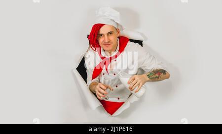 Mann mit roten Dreadlocks hält sie in seiner Handmilch. Selbstbewusster junger Koch in weißer Schürze und roter Krawatte gießt Milch aus einer Flasche in ein Glas und guckt dabei Stockfoto