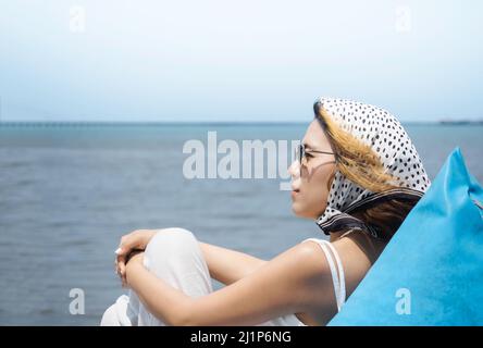 Schöne asiatische Frau Porträt in lässigen weißen Hemd tragen Sonnenbrille und Haarschal sitzen auf blauen Sitzsack am Strand mit Meerblick und blauen s Stockfoto