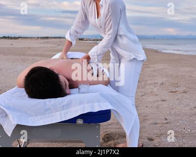 Ein junger Mann, der auf einem Massageliegen liegt und eine Chiromassage am Strand von Valencia von einem nicht erkennbaren jungen Chiromassage-Therapeuten erhält. Stockfoto
