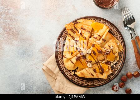 Crepes-Suzette mit Orangen und Schokolade. Dünne Crepes mit Schokoladenaufstrich, Haselnüssen und Orangenscheiben Obst auf dem Teller zum Frühstück auf altem Beton Stockfoto