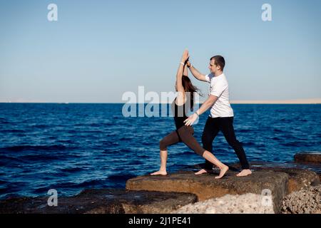 Frau Klientin tut Stretching oder Yoga im Freien Hintergrund Meer, Personal Trainer Mann korrigieren sie. Stockfoto