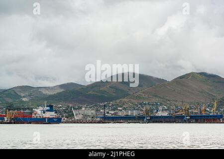 Russland, Noworossijsk, 13. August 2021 – an einem bewölkten Sommertag stehen Frachtschiffe und Hafenkrane vor der Kulisse der Berge im Hafen. Industr Stockfoto