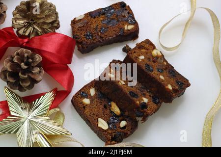 Scheiben von reichhaltigem Pflaumenkuchen mit in Rum getränkten Früchten. Garniert mit Rum getränkten getrockneten Rosinen und Preiselbeeren. Gedreht mit festlichen Dekorationen Stockfoto