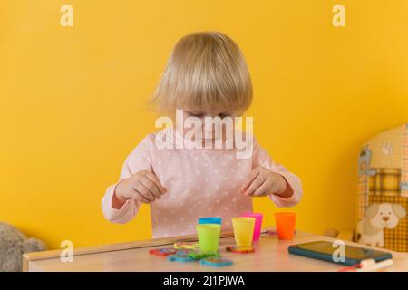 Ein Mädchen im pinken Pyjama an einem Tisch an einer gelben Wand spielt dabei, Kugeln in Tassen zu sortieren. Entwicklung der motorischen Fähigkeiten, Aufmerksamkeit, Koordination, perc Stockfoto