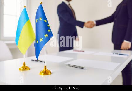 Flaggen der Ukraine und der EU vor dem Hintergrund, dass sich Vertreter von Ländern die Hände schütteln. Stockfoto