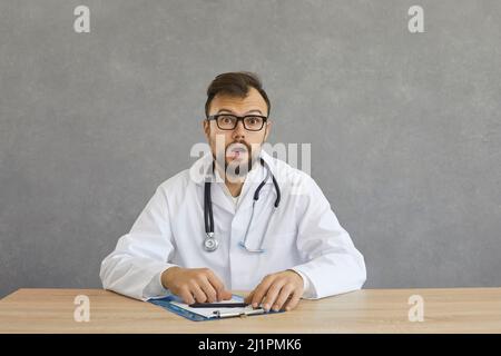 Schockierter Arzt, der am Schreibtisch sitzt und auf das Portrait des Fotostudios schaut Stockfoto