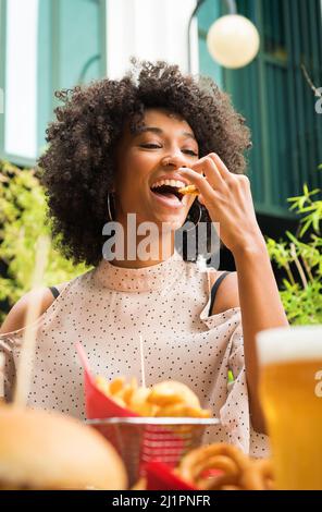 Lächelnd schöne junge glückliche schwarze Frau mit Nasenpiercing, die Kartoffelkeile in einem Pub in einem niedrigen Winkel über Essen und Gläser Bier isst Stockfoto