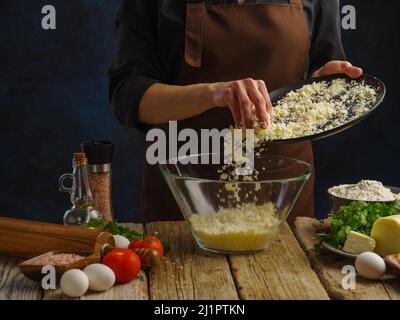 Kochen von Gerichten mit geriebenem Käse durch die Hände eines professionellen Küchenchefs auf dunkelblauem Hintergrund. Viele Produkte auf einem Holztisch. Restaurant, Hotel Stockfoto