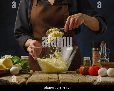 Der Koch rührt geriebenen Käse in einer Glasschüssel mit einem Löffel. Gemüse, Eier, Kräuter, Gewürze auf einem Holztisch. Dunkler Hintergrund. Kochen von Gerichten mit GRA Stockfoto