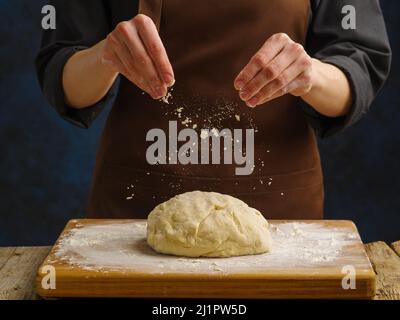 Zubereitung von Teig durch die Hände eines professionellen Küchenchefs auf einem Holzschneidebrett auf dunklem Hintergrund. Der Koch streut den Teig mit Mehl. Levi Stockfoto