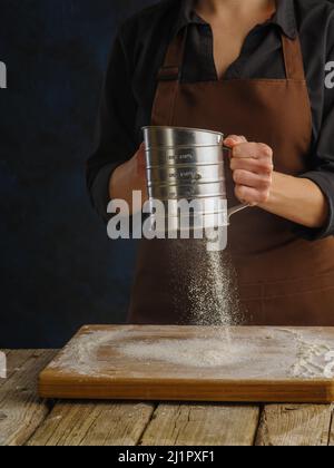 Der Prozess der Zubereitung von Teig mit den Händen des Küchenchefs auf einem dunklen Hintergrund. Der Koch siebt das Mehl durch ein Sieb. Levitation. Nahaufnahme. Das Konzept Stockfoto