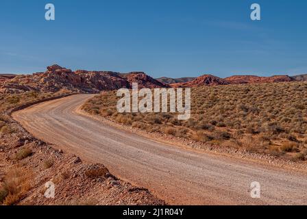 Overton, Nevada, USA - 11. Dezember 2010: Valley of Fire. Graue Schotterstraße macht breite Wendung auf trockenem Wüstenboden voll von grünen Büschen unter blauem Himmel und r Stockfoto