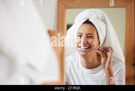 Das Zimmer leuchtet buchstäblich auf, wenn sie lächelt. Aufnahme einer jungen Frau, die sich die Zähne putzt, während sie in den Badezimmerspiegel schaut. Stockfoto