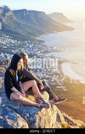 Genießen Sie die Aussicht von oben. Ganzkörperaufnahme eines jungen Paares, das auf einem Berggipfel sitzt. Stockfoto