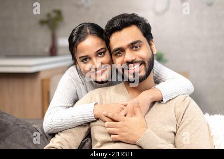 Porträt von schönen Paar zu Hause. Schöner Mann, der auf der bequemen Couch sitzt, schöne Frau, die sich von hinten umarmt. Ehepartner verbringen Zeit miteinander Stockfoto