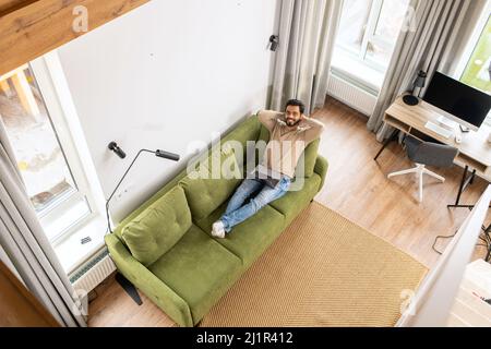 Fröhlicher inder in legerer Kleidung liegt auf dem bequemen Sofa und nutzt einen trendigen Laptop für Videoanruf, lächelt und entspannt Stockfoto