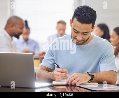Entschlossenes Arbeiten in einem stark frequentierten Büro. Aufnahme eines jungen Geschäftsmannes, der sich Notizen machte, während er mit seinen Kollegen in einem Büro an einem Laptop arbeitete Stockfoto