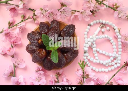 Dattefrüchte in Holzplatte und weißen Gebetsperlen isoliert auf rosa Hintergrund. Ramadhan Konzept Idee. Blumenrahmen. Nahaufnahme, Draufsicht. Stockfoto