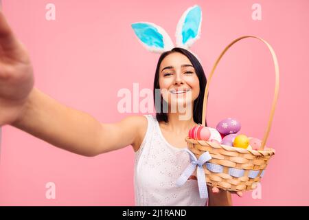 Schöne fröhliche Frau in Kaninchenohren macht ein Selfie, hält einen Korb mit Yayyuami in den Händen, Videoanruf, ostern, im Studio auf einem rosa Backgro Stockfoto