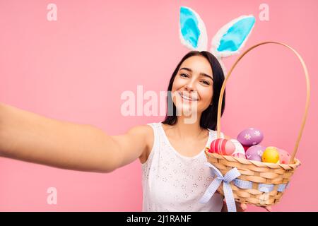 Schöne fröhliche Frau in Kaninchenohren macht ein Selfie, hält einen Korb mit Yayyuami in den Händen, Videoanruf, ostern, im Studio auf einem rosa Backgro Stockfoto