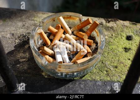 Eine große Anzahl von Zigarettenenden, die in einem runden Glasaschenbecher außen an einer Wand stehen Stockfoto