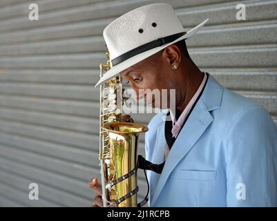 Der hübsche, elegante junge, lustige afro-kubanische Saxophonist trägt einen weißen panamahut und blickt in sein Saxophon hinab. Stockfoto