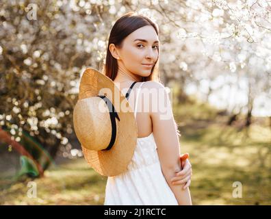 Mädchen mit einem Strohhut im Frühling im Park. Brunette mit langen Haaren hält einen Hut auf einem Hintergrund der Sommernatur. Jugend und Schönheit. Stockfoto