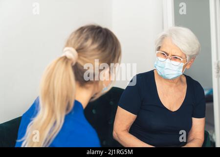 Blonde Krankenschwester in blauer Uniform hat einen Hausbesuch. Ältere maskierte Dame, die sich über gesundheitliche Probleme beschwert. Hochwertige Fotos Stockfoto