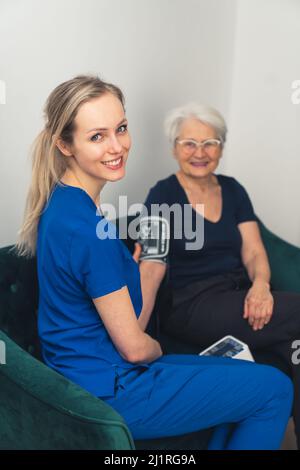 Sich um das eigene Gesundheitskonzept kümmern. Die ältere Oma und ihre schöne junge Pflegekraft sitzen während ihres Hausbesuches auf einem Sofa zusammen. Hochwertige Fotos Stockfoto