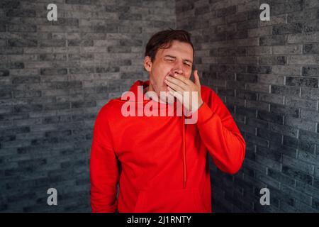 Mann in einem roten Hoodie gähnt vor Müdigkeit auf einem Backstein-Hintergrund. Stockfoto