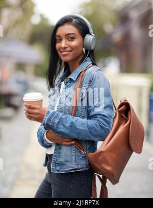 Kaffee hält mich in Gang. Aufnahme einer Frau, die Kopfhörer trägt und draußen einen Kaffee zum Mitnehmen hält. Stockfoto