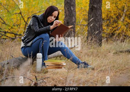 Eine junge, schöne Frau mit langen, dunklen Haaren in legerer Kleidung sitzt auf einem Holzbuch und liest ein Buch Stockfoto