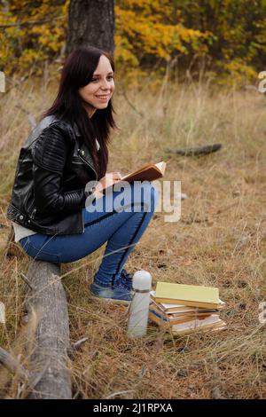 Eine junge, schöne Frau mit langen, dunklen Haaren in legerer Kleidung sitzt auf einem Holzbuch und liest ein Buch Stockfoto