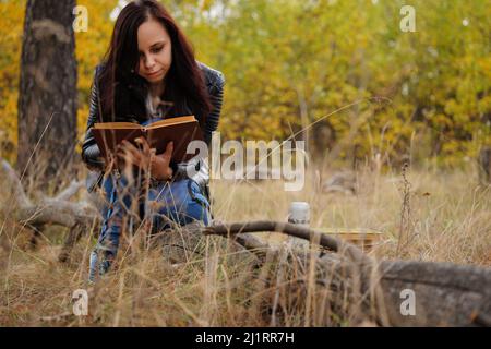Eine junge, schöne Frau mit langen, dunklen Haaren in legerer Kleidung sitzt auf einem Holzbuch und liest ein Buch Stockfoto