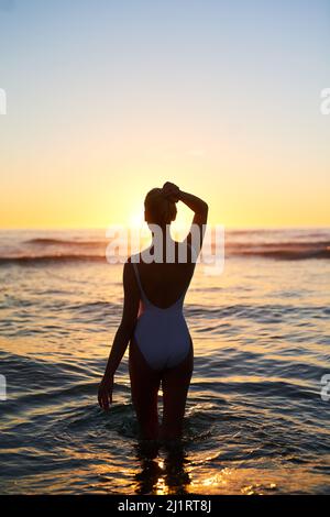 Wir alle verlieren uns manchmal in der Schönheit. Rückansicht einer jungen Frau, die bei Sonnenuntergang im Wasser steht. Stockfoto