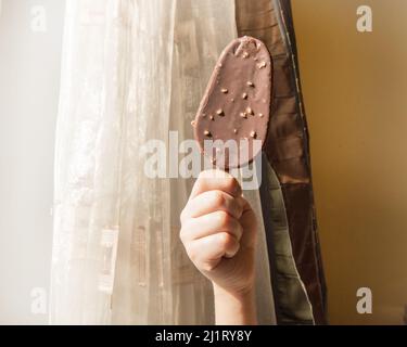 Eine Kinderhand hält ein Schokoladeneis auf einem Stock, ein Eis mit Nüssen auf dem Hintergrund eines Wohninnenraums an einem sonnigen Tag. Stockfoto