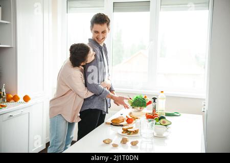 Liebevolle Frau umarmt ihren Freund von hinten Zeit in der Küche zu verbringen. Vegetarisch gesunde Lebensmittel. Aktivitätsbeziehung. Kochen, zusammen, Küche Stockfoto
