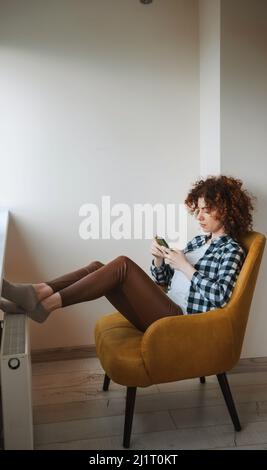 Lockige Frau, die auf einem Sessel sitzt und online mit ihrem Mobiltelefon spricht. Über Telefon. Porträt einer Geschäftsfrau. Mobile Kommunikation. Stockfoto