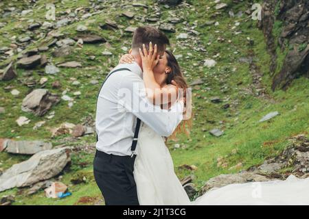 Bräute umarmen und küssen, während sie für ein Foto auf dem felsigen Berg Hügel Hintergrund posieren. Hochzeitsreise. Wunderschöne Sommerlandschaft. Stockfoto